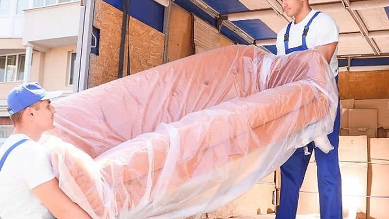 Dos operarios de mudanzas cargando un sofá embalado en film dentro del camión